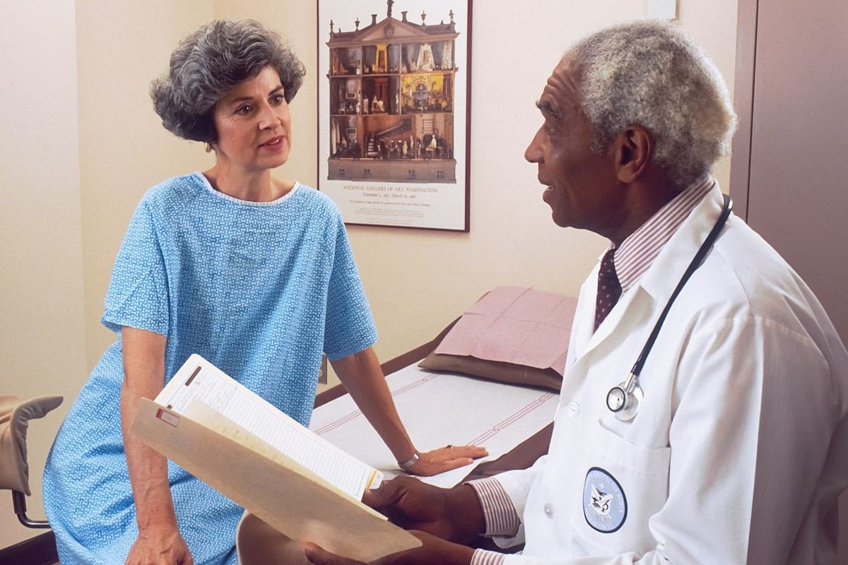 An older doctor talking with a patient seated on an examination couch.