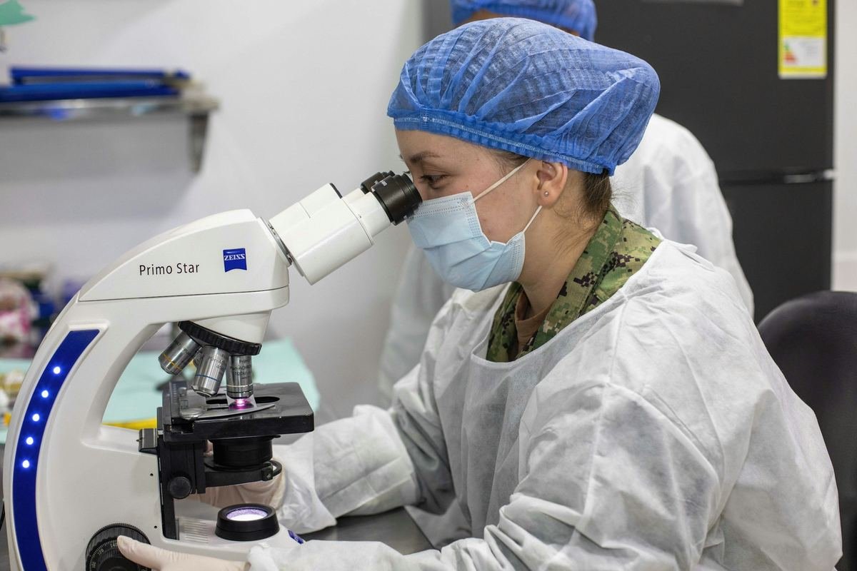A laboratory scientist in a cap and mask examining a sample under a microscope.